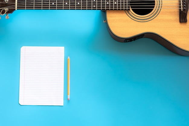 Acoustic guitar and blank paper on blue background, top view, concept of musical creativity, hobby.