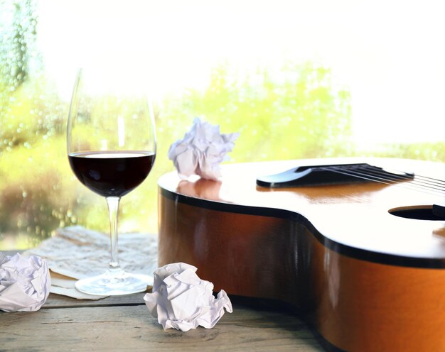 雨滴のある窓の隣にアコースティックギターとグラスワイン