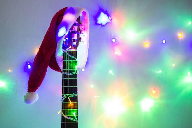 Acoustic guitar with red Santa hat and light colorful garland. Christmas music concept on white background with copyspace