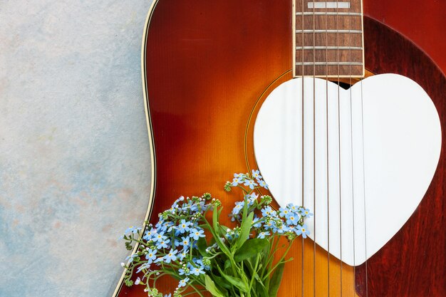 Acoustic guitar, wooden white heart shape symbol and forget-me-not flowers on sky blue background. Top view, close up, copy space.