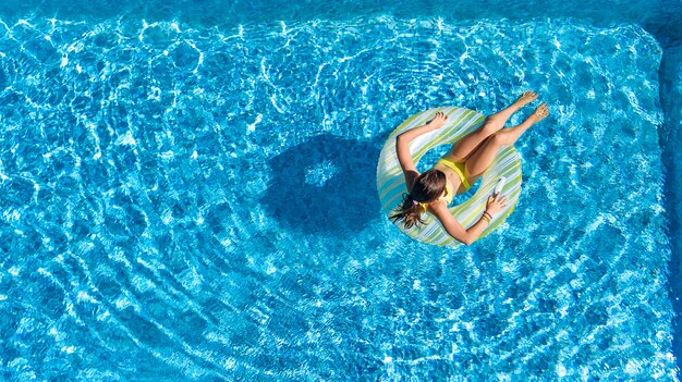 Acrive girl in swimming pool aerial top view from above, kid swims on inflatable ring donut , child has fun in blue water on family vacation resort