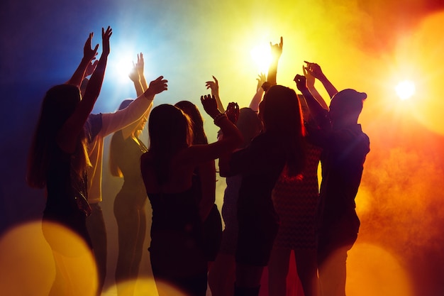 Actie. Een menigte van mensen in silhouet steekt hun handen op dansvloer op neonlichtachtergrond. Nachtleven, club, muziek, dans, beweging, jeugd. Geelblauwe kleuren en bewegende meisjes en jongens.