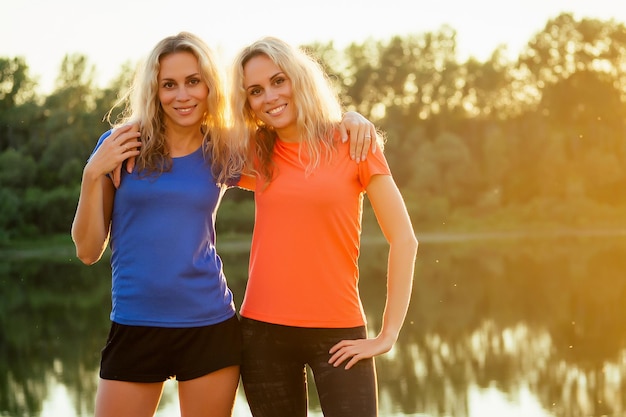 Actieve familie op buiten. twee flexibiliteit krullend blonde mooie vrouw tweelingzusjes in stijlvolle sportkleding het lichaam uitrekken en warming-up joggen in het stadion meer mooie zonsondergang achtergrond park.