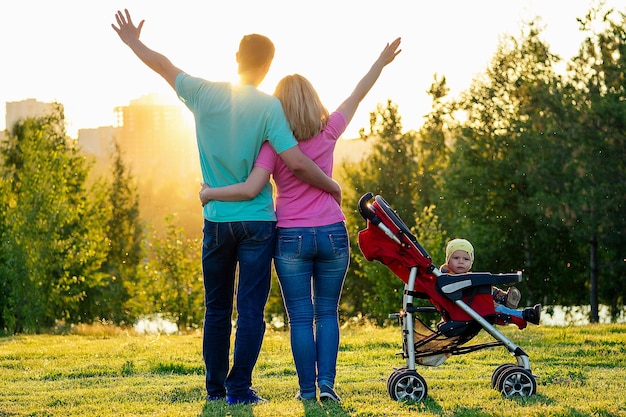 Actieve gelukkige familie die in het zomerpark loopt met een rode kinderwagen met dochterzoon, ze strekten hun armen uit als het vrijheidsconcept van vogels