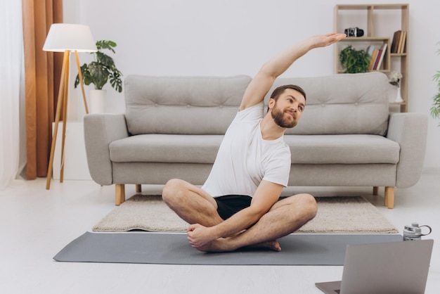 Actieve jonge vrolijke man die ochtendgymnastiek doet thuis zittend op yogamat en uitrekkende kopieerruimte