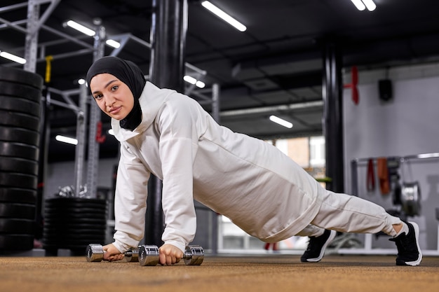 Actieve moslimvrouw in hijab met intensieve training die push-ups plank met halters doet. crossfit-oefeningen bij gymnastiek, sportconcept