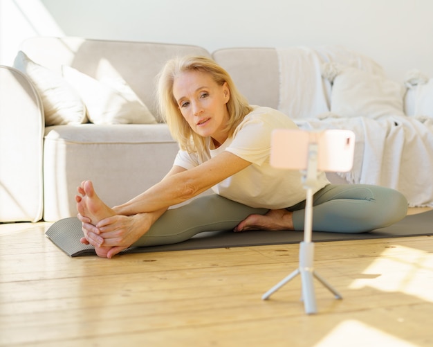 Actieve senior vrouw die traint op yogamat in de woonkamer tijdens het kijken naar online les op smartphone