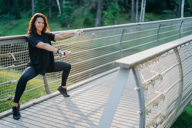 Actieve vastberaden vrouw doet squat oefening met halters poses in actieve slijtage bij brug maakt gebruik van sportuitrusting verhoogt gewicht en traint biceps heeft blije uitdrukking Sportief levensstijl concept