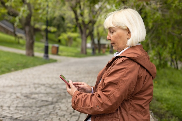 Actieve volwassen senior vrouw gebruikt smartphone terwijl ze op een bankje in het park zit