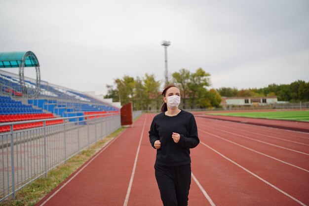 Actieve vrouw met beschermend masker die in het stadion loopt Jonge fitte vrouw in sportkleding en beschermend masker voor coronaviruspreventie die op rode baan loopt tijdens buitentraining in het stadion