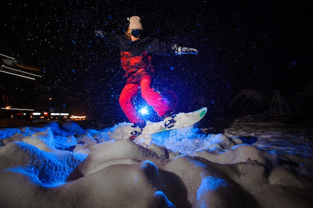 Actieve vrouwelijke snowboarder gekleed in een rode sportkleding die 's nachts op de berghelling springt onder het blauwe licht