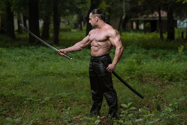 Action Hero Muscled Man Holding A Ancient Sword  Standing In Forest Wearing Leather Pants