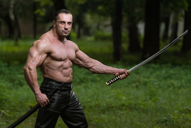 Action hero muscled man holding a ancient sword standing in\
forest wearing leather pants