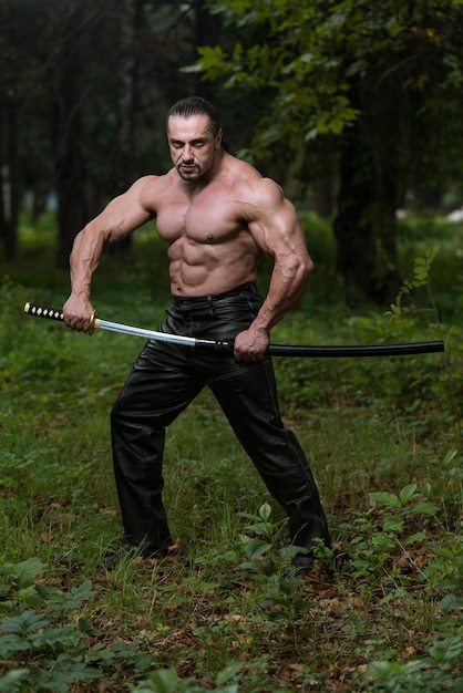 Action Hero Muscled Man Holding A Ancient Sword  Standing In Forest Wearing Leather Pants