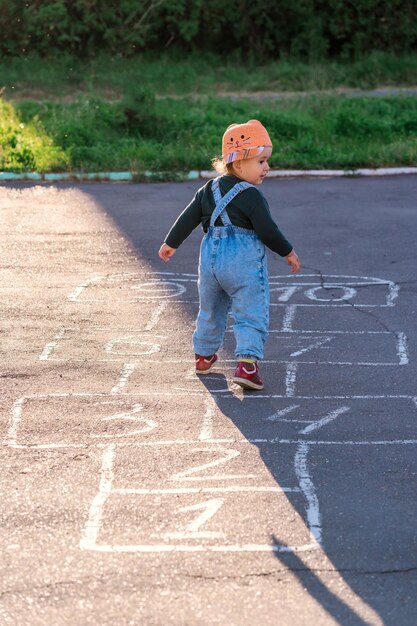 アクティブな 2 歳の女の子がアスファルトにペイントされた石けり遊びを学ぶ