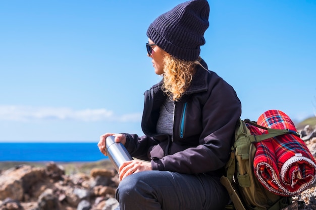 Active adult lady resting and taking a break while do trekking
adventure activity inthe nature. woman sit down on the rocks
admiring ocean blue view. people travel with backpack