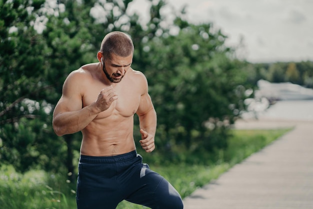 Active bearded man runs outdoors listens to music has a muscular body poses near trees