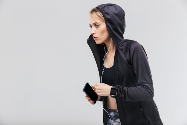 активная белокурая женщина, одетая в спортивную одежду, слушает музыку в наушниках во время бега, изолирована над белой стеной