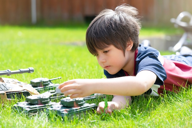Active boy laying down on the grass playing with soldiers and tank toys in the garden