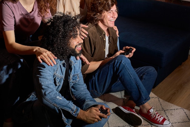 Active couples friends enjoying playing videogame on game\
console at home holding gamepad in hands, guys on floor engaged in\
game while their girlfriends supporting them