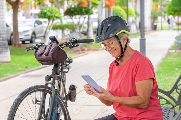 Donna anziana ciclista attiva seduta su una panchina nel parco pubblico che legge un messaggio sul telefono cellulare