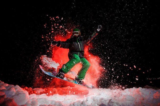 사진 붉은 빛 아래에서 밤에 눈 아래에서 점프하는 녹색 운동복을 입은 활성 여성 스노