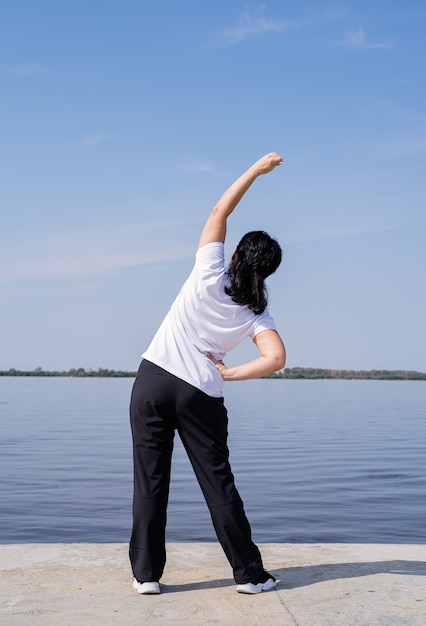 Active and happy senior woman doing stretching near the riverside