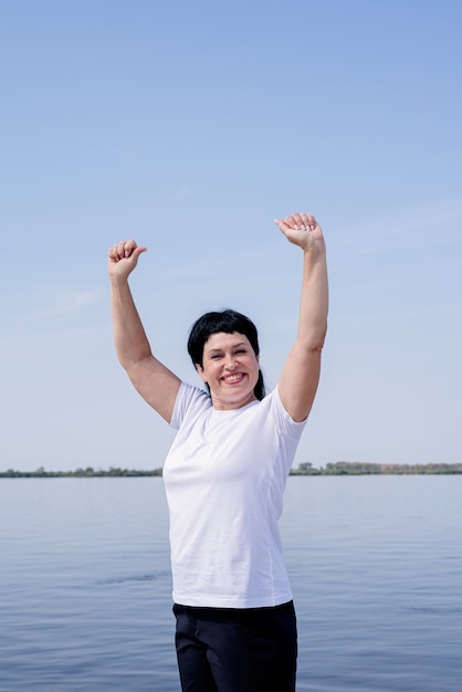 Active and happy senior woman exercising near the riverside standing with arms up