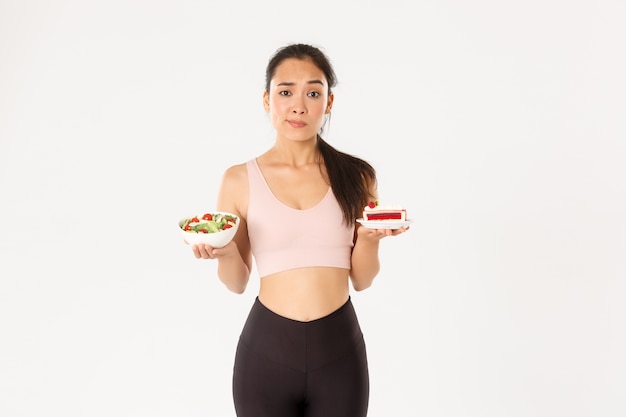 Active lifestyle, fitness and wellbeing concept. Indecisive cute asian slim girl in workout outfit, holding healthy salad and cake, hesitating, need stay on diet, losing weight 