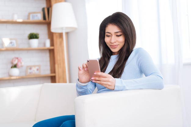 Active mailing. Thoughtful pretty woman smiling while holding smartphone