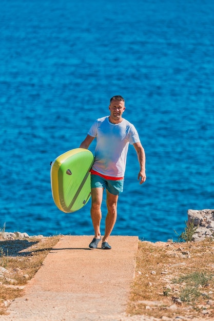 Foto uomo attivo che porta una tavola da paddle colorata su un sentiero costiero soleggiato