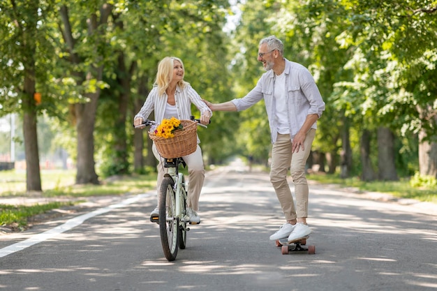 Active mature couple riding bike and skateboard on path in park