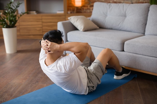 Active mature man exercising abs muscles on yoga mat near sofa at home doing domestic fitness