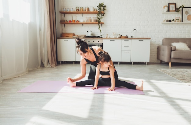 An active mother and a little daughter in black suits are\
engaged in fitness yoga physical exercises at home sports\
family
