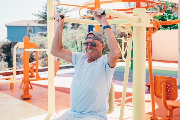 Active old senior man doing exercises on public open air gym equipment looking smiling at camera