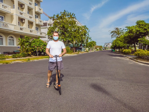 Active outdoor life in surgical sterilizing face mask Young beautiful man riding on adult scooter in modern cottage village Protecting from air pollution and virus infection as new normal lifestyle