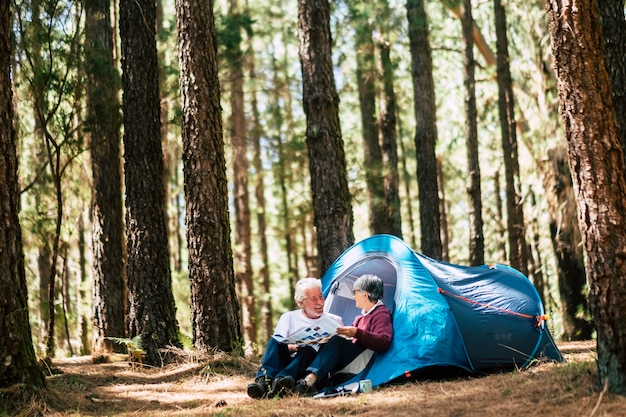 Active people old aged caucasian senior couple travel with camping tent - sit down in the forest having fun together - relationship forever concept for traveler wanderlust retired man and woman