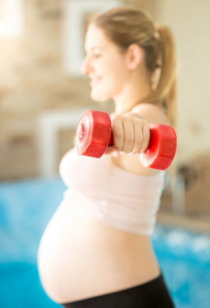 Active pregnant woman doing exercise with dumbbells at fitness club. Side view, focus on hand.