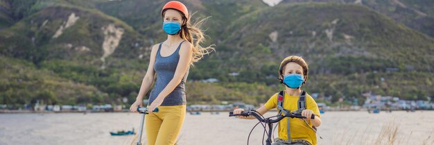医療用マスクと安全ヘルメットをかぶった活発な学童の少年とその母親が、バックパックを背負って自転車に乗っている