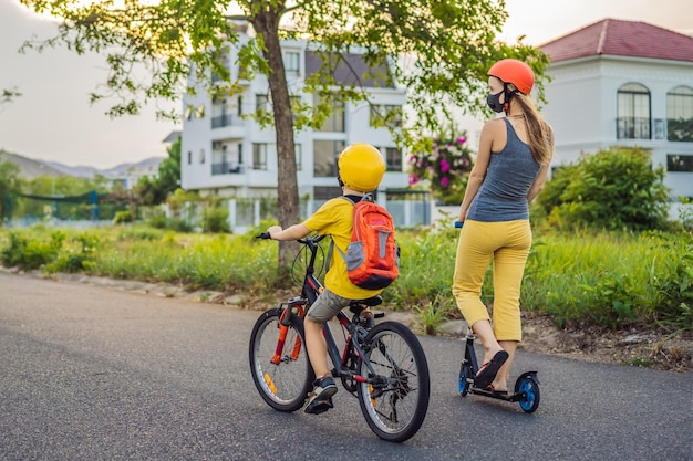 Active school kid boy and his mom in medical mask and safety helmet riding a bike with backpack on