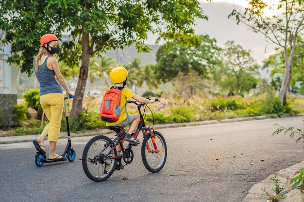 医療用マスクと安全ヘルメットをかぶった活発な学童の少年とその母親が、バックパックを背負って自転車に乗っている