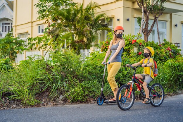 Active school kid boy and his mom in medical mask and safety helmet riding a bike with backpack on