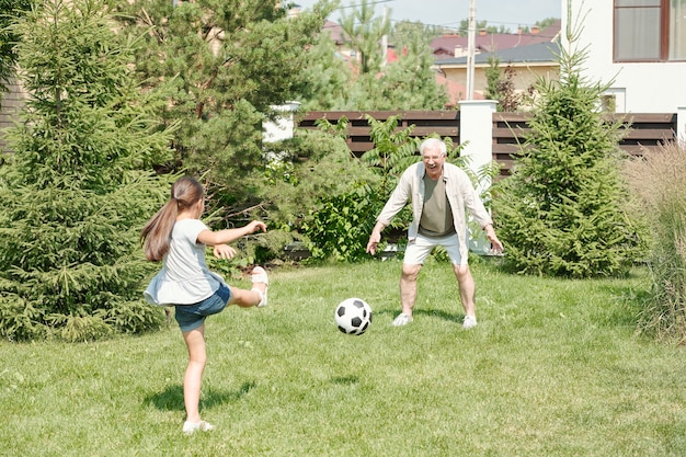 Active senior man playing soccer with his little granddaughter on lawn in backyard