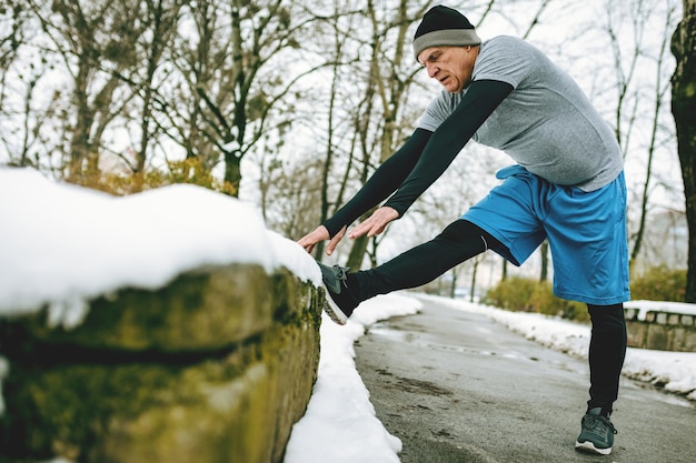 Active senior man stretching and doing exercises in public park during the winter training outside in.