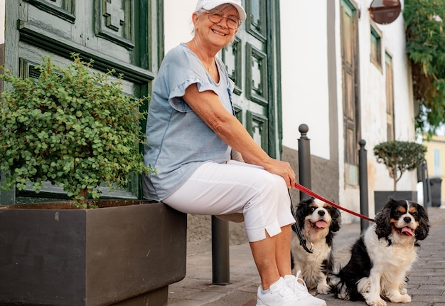 Active senior smiling woman with hat sitting in the street with her two cavalier king Charles dogs