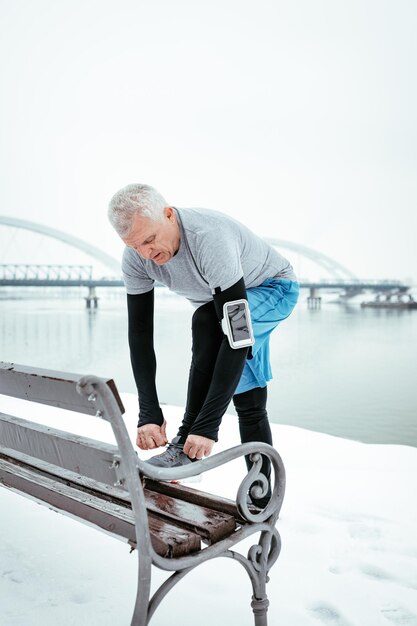 Active senior tying running shoes, and making pause during stretching and doing exercises by the river during the winter training outside in. Copy space.