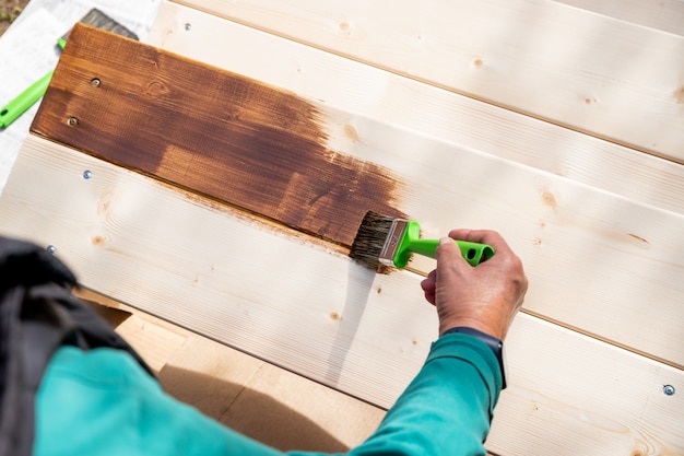 Active senior woman painting some pieces of wood, timber by brown paint colour with brush.worker painting a wooden wall