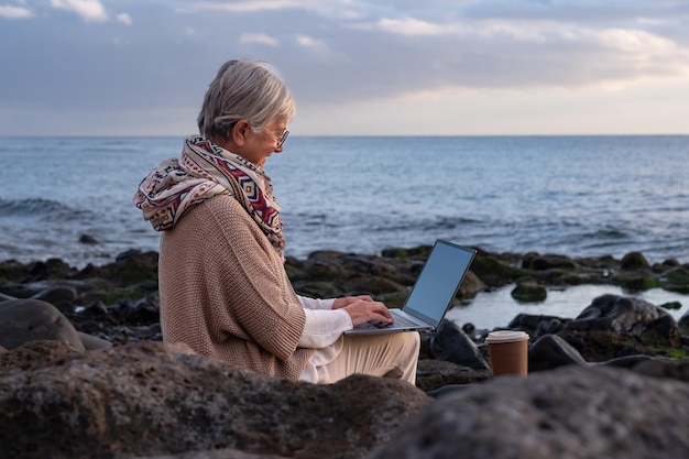 Active senior woman sitting on the rocks beach using laptop Elderly caucasian female in online remote work enjoying sunset at sea horizon over water