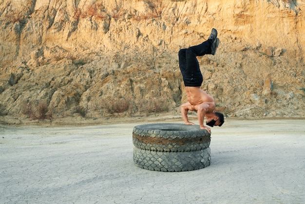 Active shirtless guy balancing on tyres and holding legs up while training at sand pit