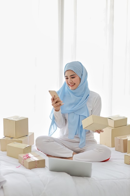 Active smiling asian muslim woman in sleepwear sitting on bed using mobile phone and computer. Startup small business SME freelance woman working with online package box delivery, E-commerce concept.